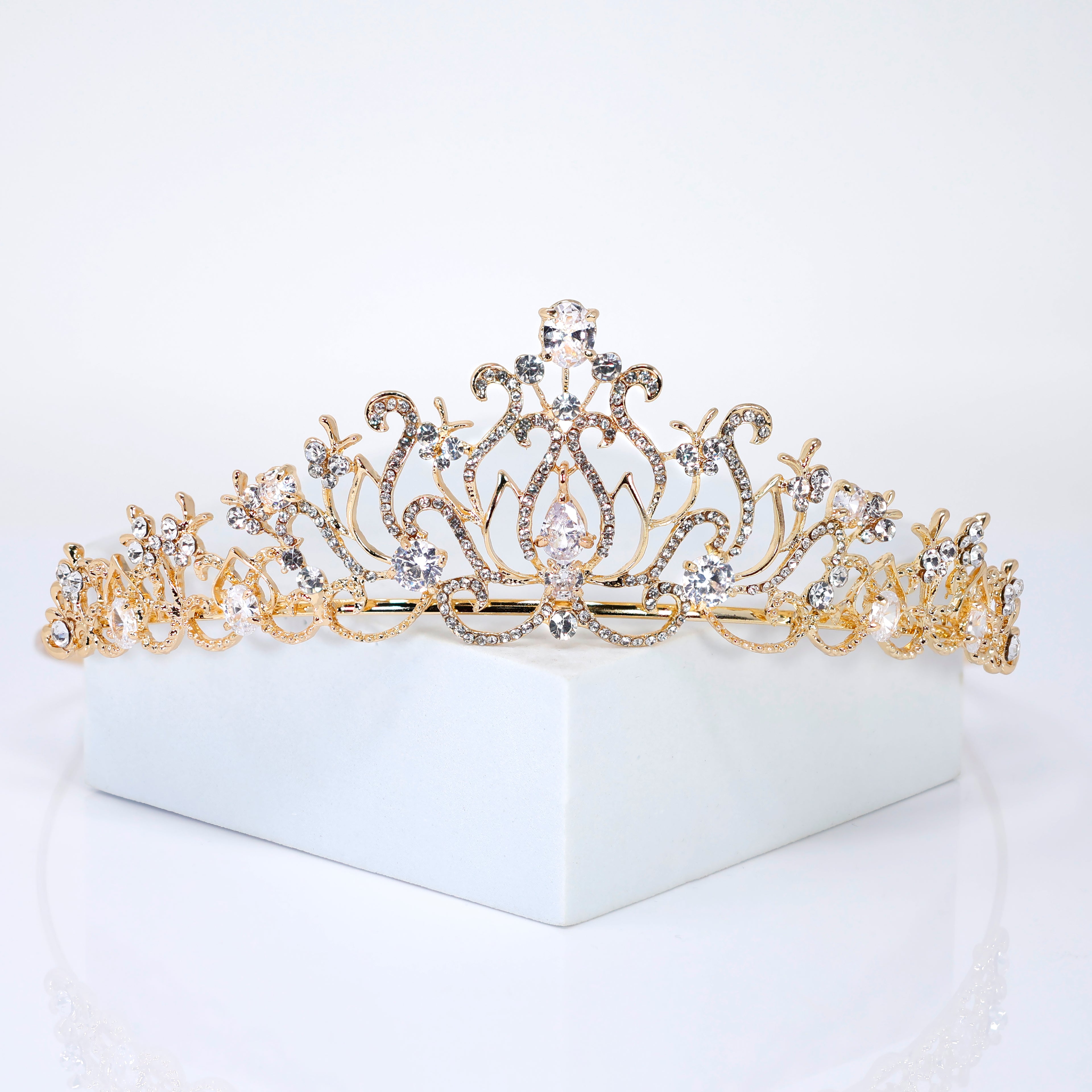 A gold tiara with a floral design and sparkling gemstones is displayed on a white surface.