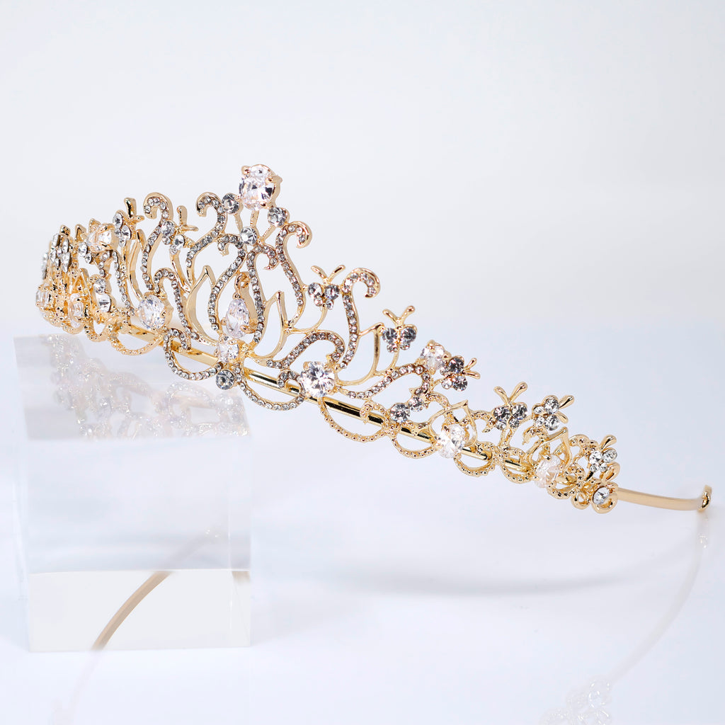 A gold tiara with intricate designs and sparkling gemstones is displayed on a clear glass stand against a white background.
