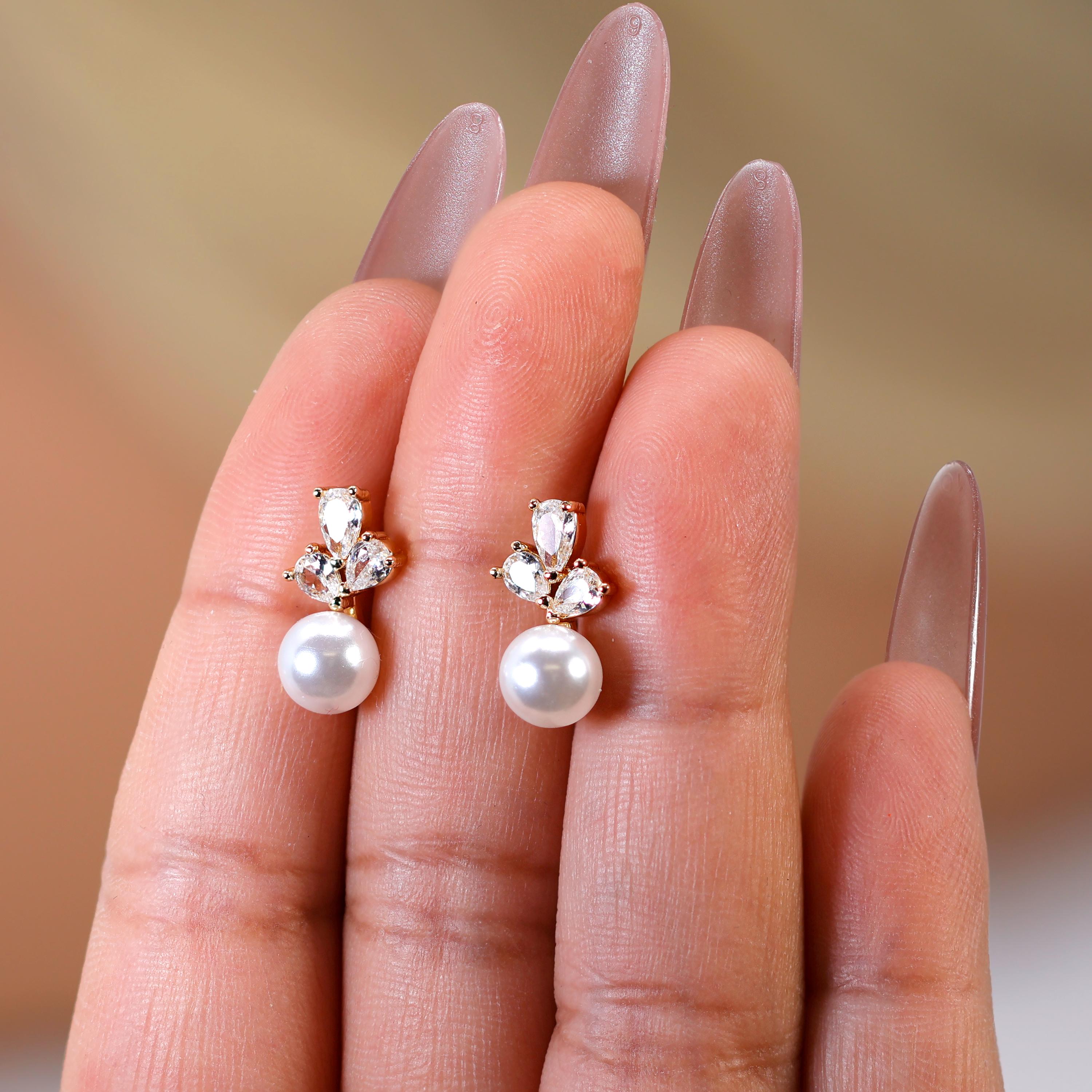 A close-up of a person's hand wearing a pair of pearl drop earrings with gemstone accents.