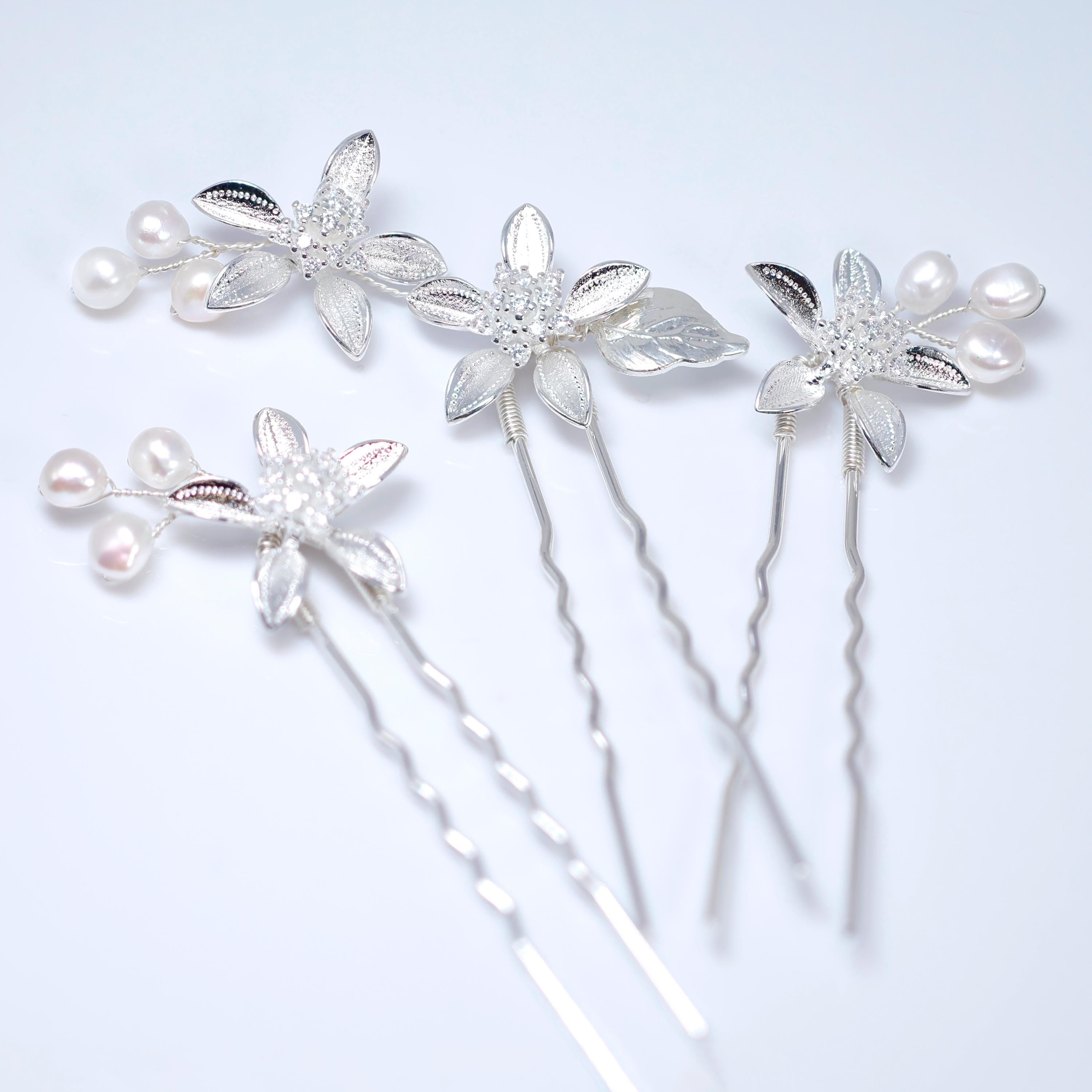 a collection of silver hair pins with flower-shaped designs, including pearls and leaves, arranged on a white background.