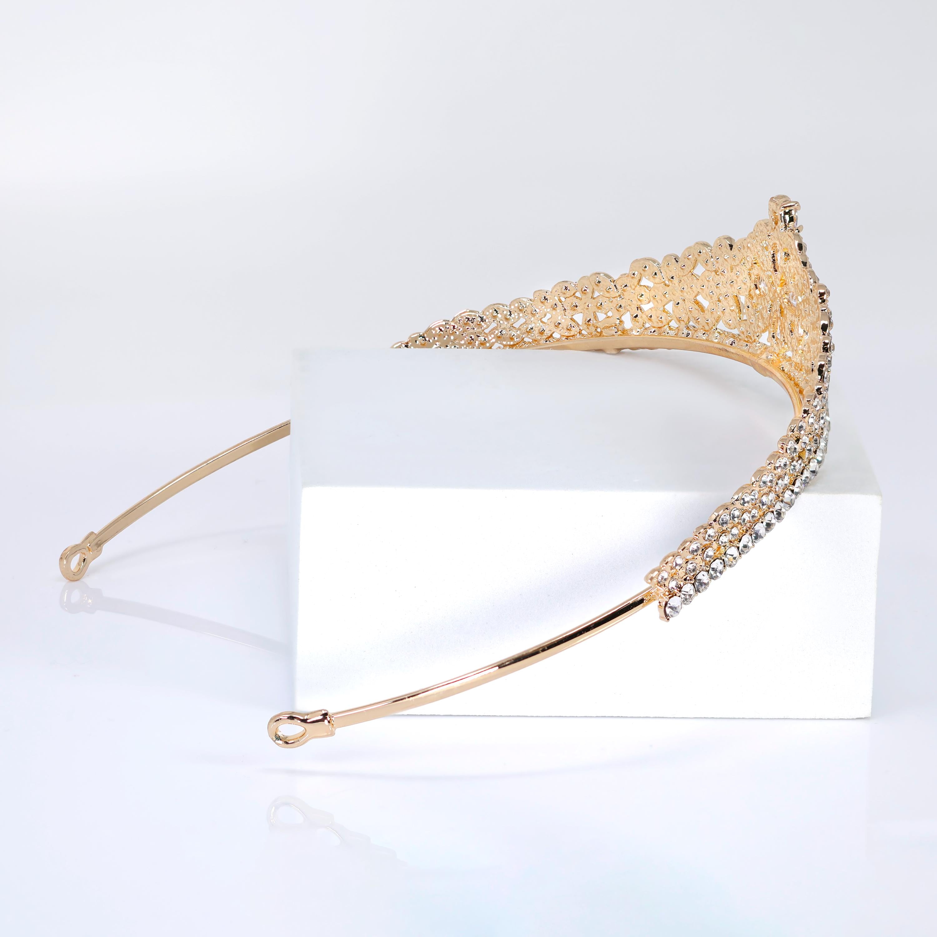A gold tiara with a diamond-encrusted design rests on a white surface.